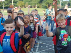 Children wearing backpacks in a line outdoors giving "thumbs up"